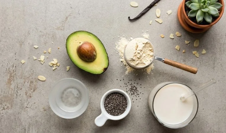 A flat-lay of protein powder, avocado, MCT oil, and almond milk, showcasing the essential ingredients used to prepare high-quality low carb meal replacement shakes.