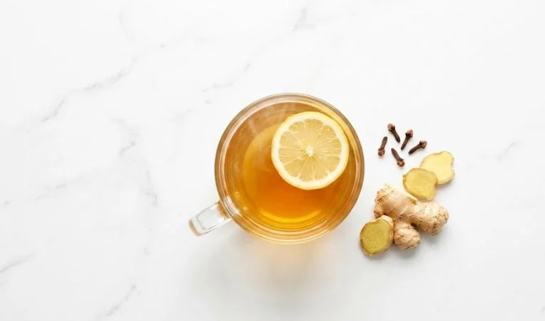 A clear glass mug of ginger and lemon cold and flu tea with a floating lemon slice, styled on a bright white marble background for a healthy lifestyle blog.